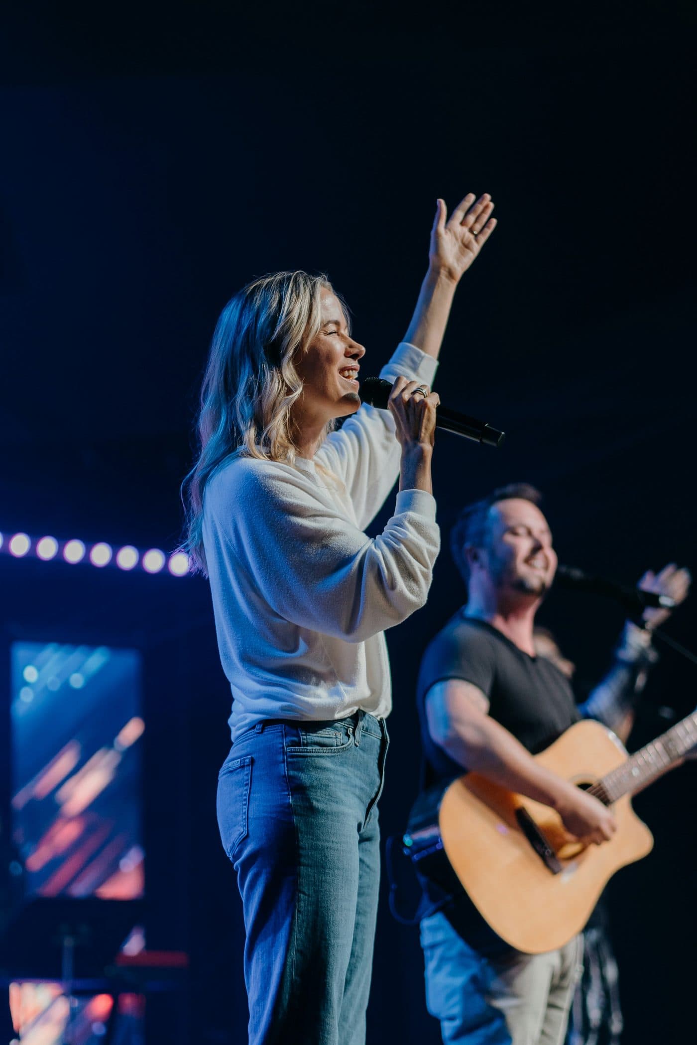 A worship leader singing on stage with a guitarist beside her.
